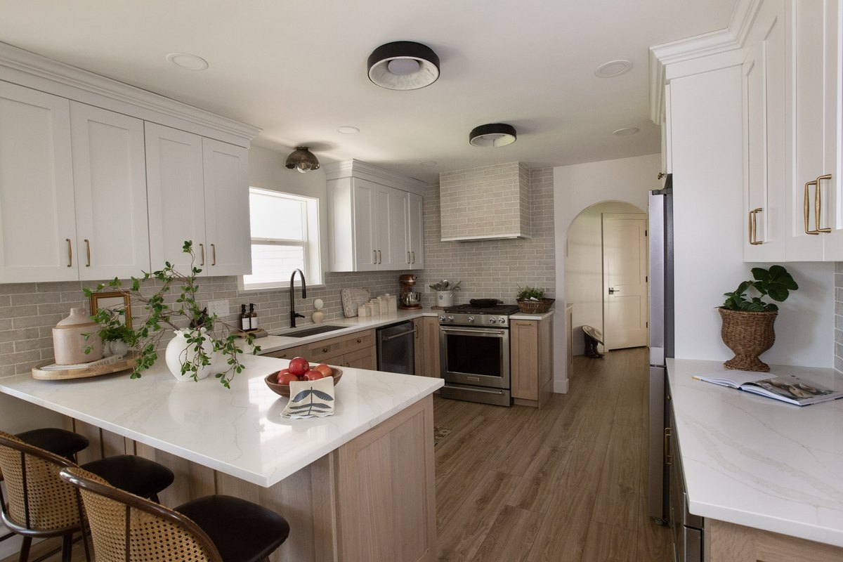 A white and oak kitchen with a marble-topped peninsula, greenery on a wooden tray, and a bowl of apples