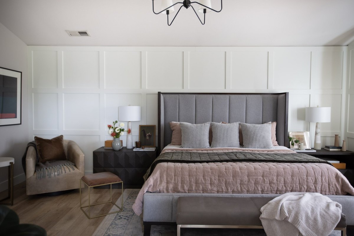 A bedroom with a panel-molded white wall, a tall gray upholstered bed layered in blush linen and an olive quilt, houndstooth pillows, and a velvet swivel chair in the corner