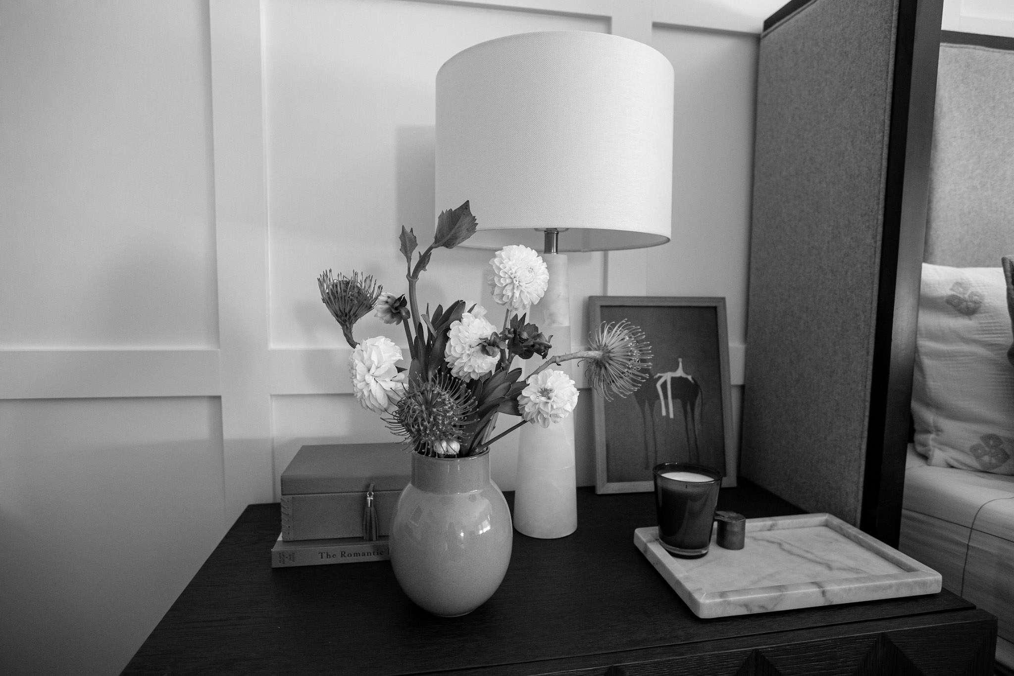 A black and white photograph of a bedside table styled with an alabaster lamp, dahlias in a ceramic vase, a framed print, and a candle on a marble tray