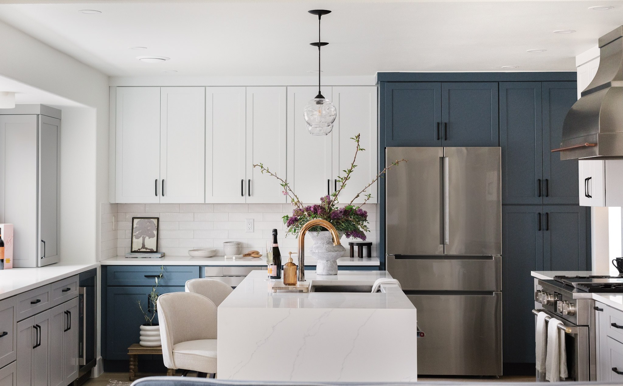 A kitchen with navy and white cabinetry, a waterfall quartz island, brass faucet, and lilacs in a stone vase