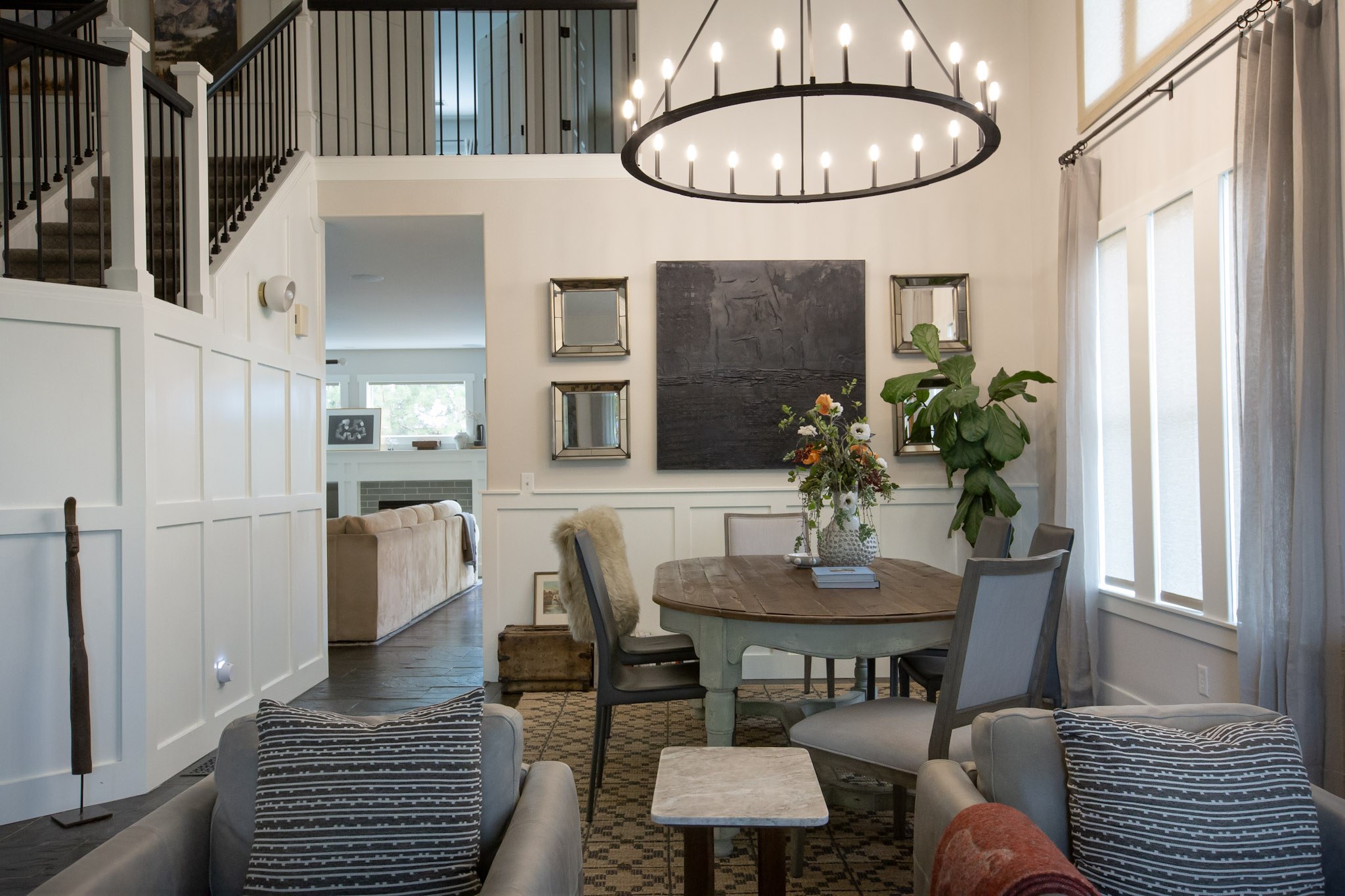 A dining area with a round farmhouse table, a large iron ring chandelier, paired mirrors, and a fiddle-leaf fig by the window