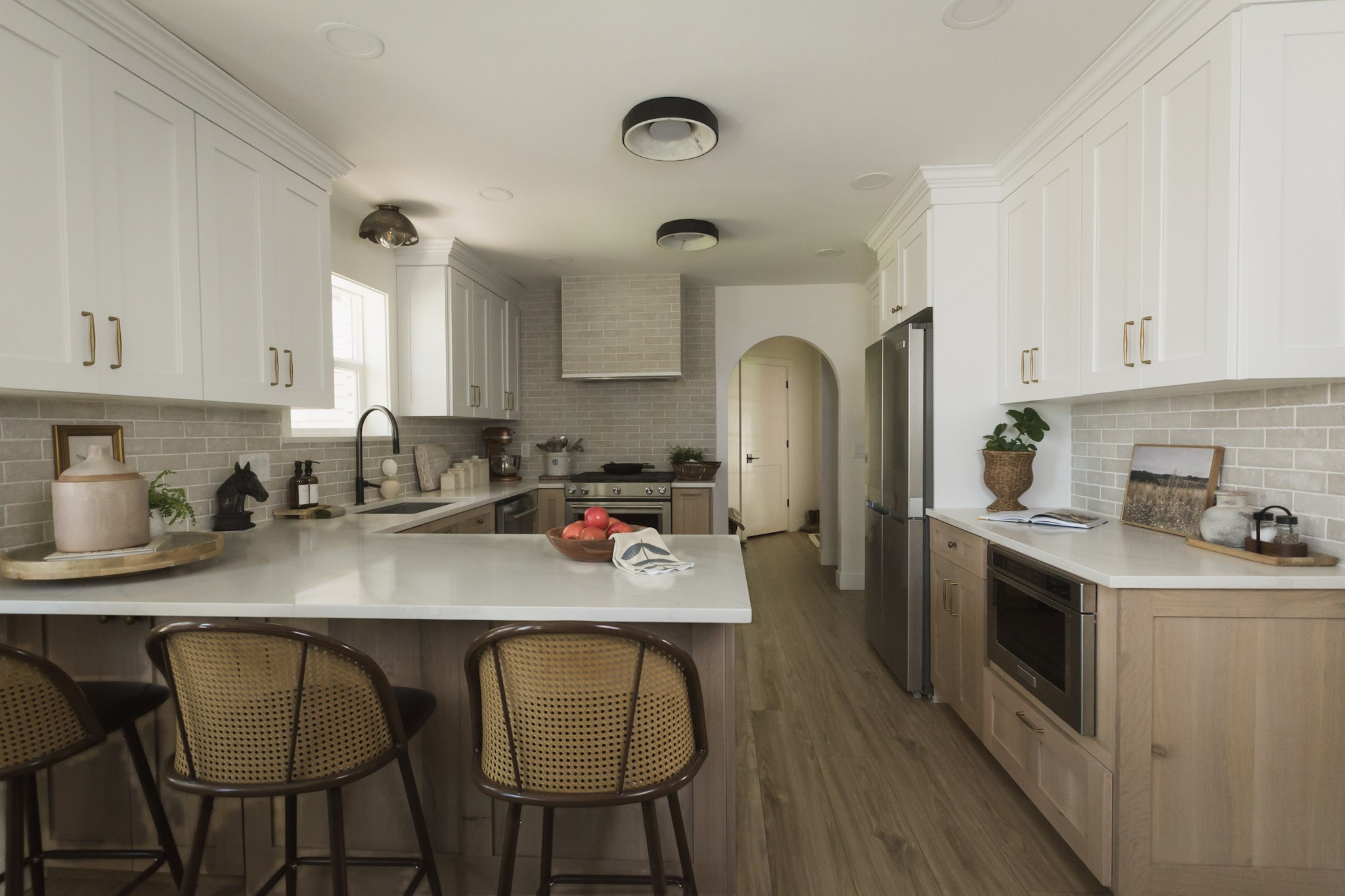 A white and oak kitchen with a marble peninsula, cane counter stools, gray tile backsplash, and an arched doorway beyond