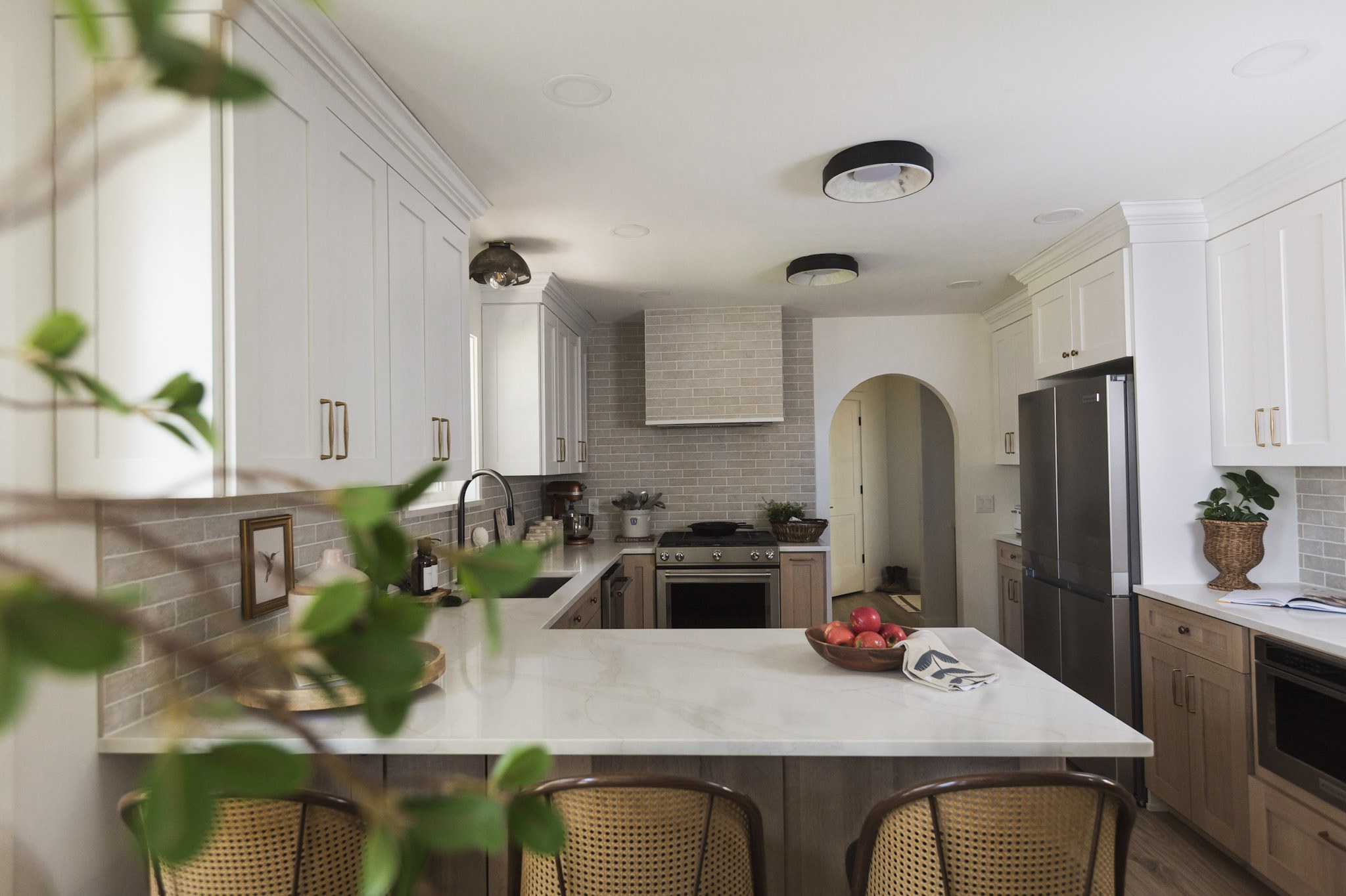 The same kitchen framed through the leaves of a potted branch, with a bowl of apples on the marble peninsula